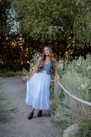 A person in a flowing light blue dress and brown boots twirls on a desert path among desert plants at sunset.