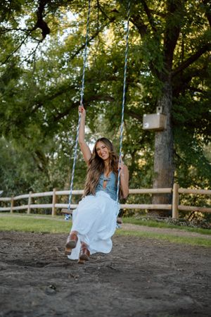 Woman in casual white top swings joyfully on outdoor tree swing during golden hour in summer backyard setting.
