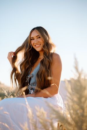 A woman in a denim top and white skirt smiles while sitting outdoors during golden hour in a field.