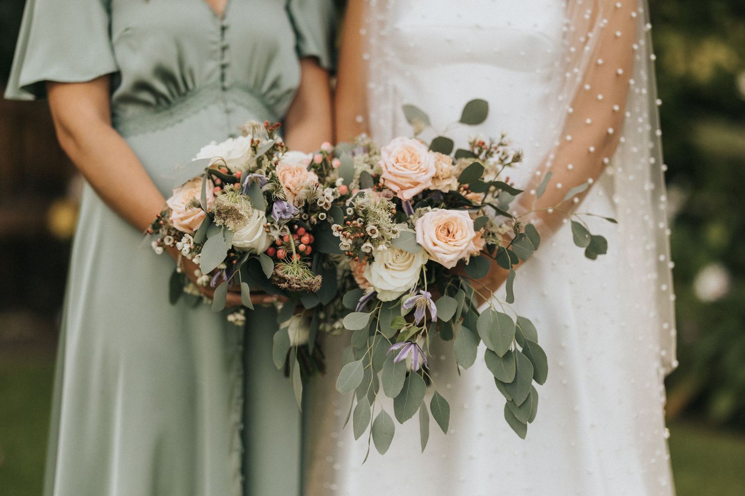 Two wedding bouquets with blush roses and eucalyptus leaves held against sage green bridesmaid and white bridal dresses.