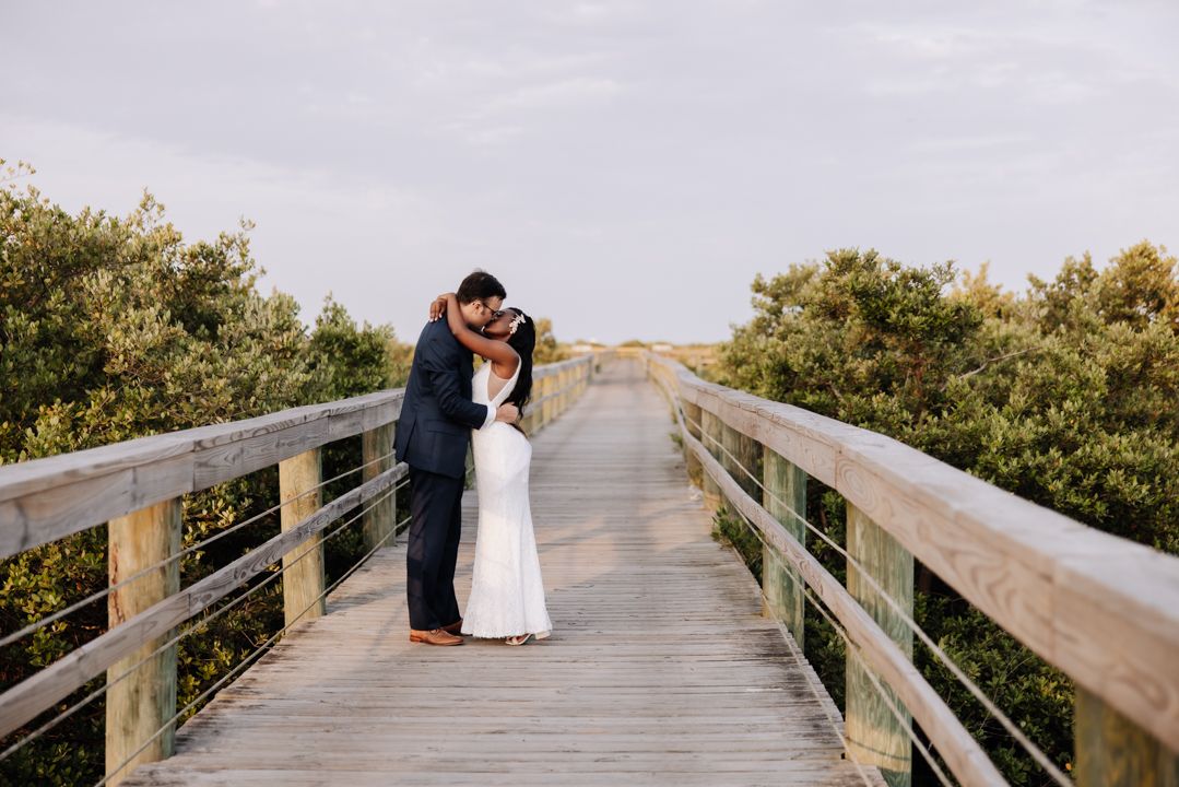 Couple poses for romantic wedding photos on wooden boardwalk surrounded by lush coastal greenery at sunset.