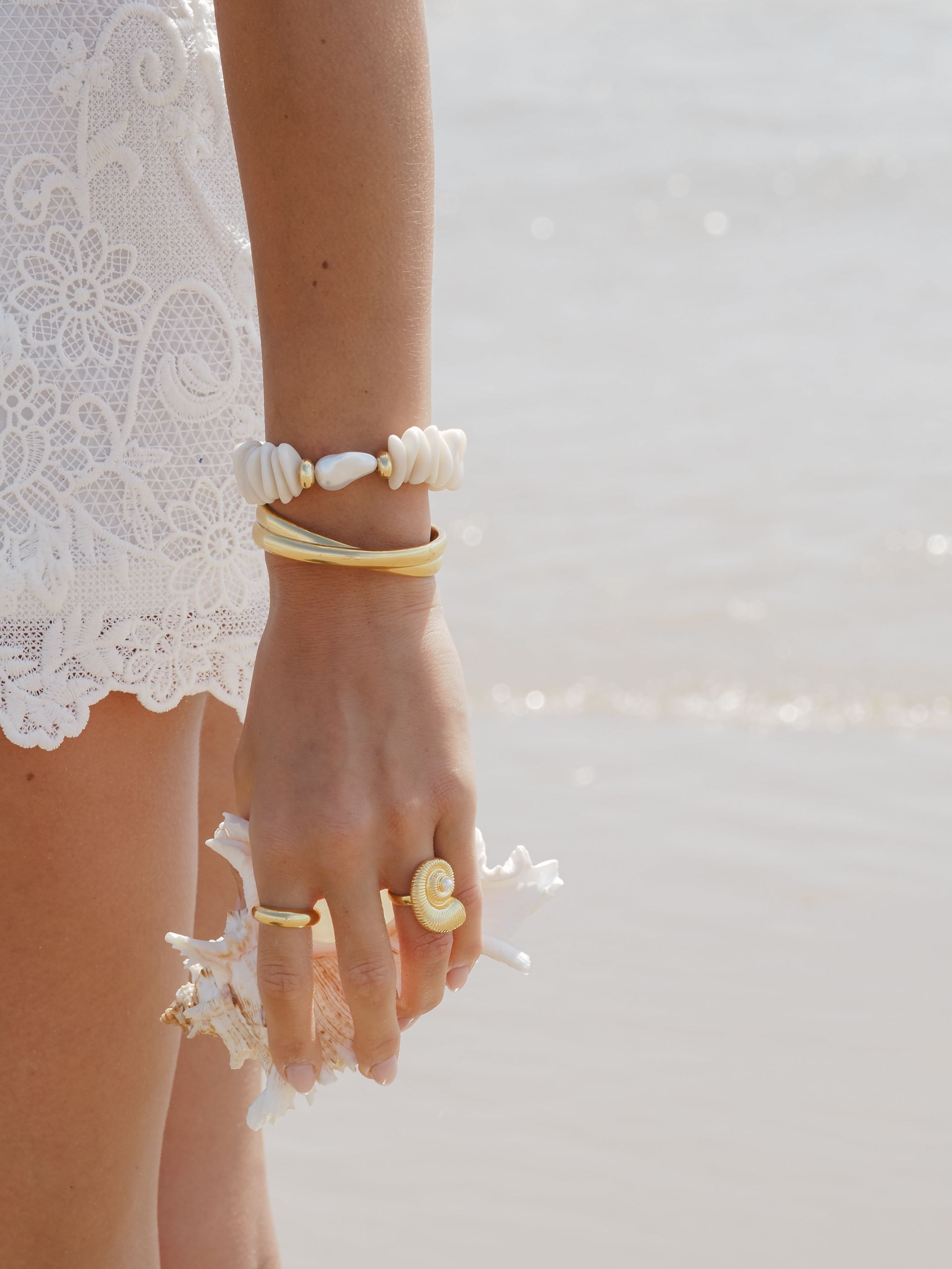 Delicate shell and seashell jewelry pieces including bracelets and rings displayed on a hand against white lace fabric.