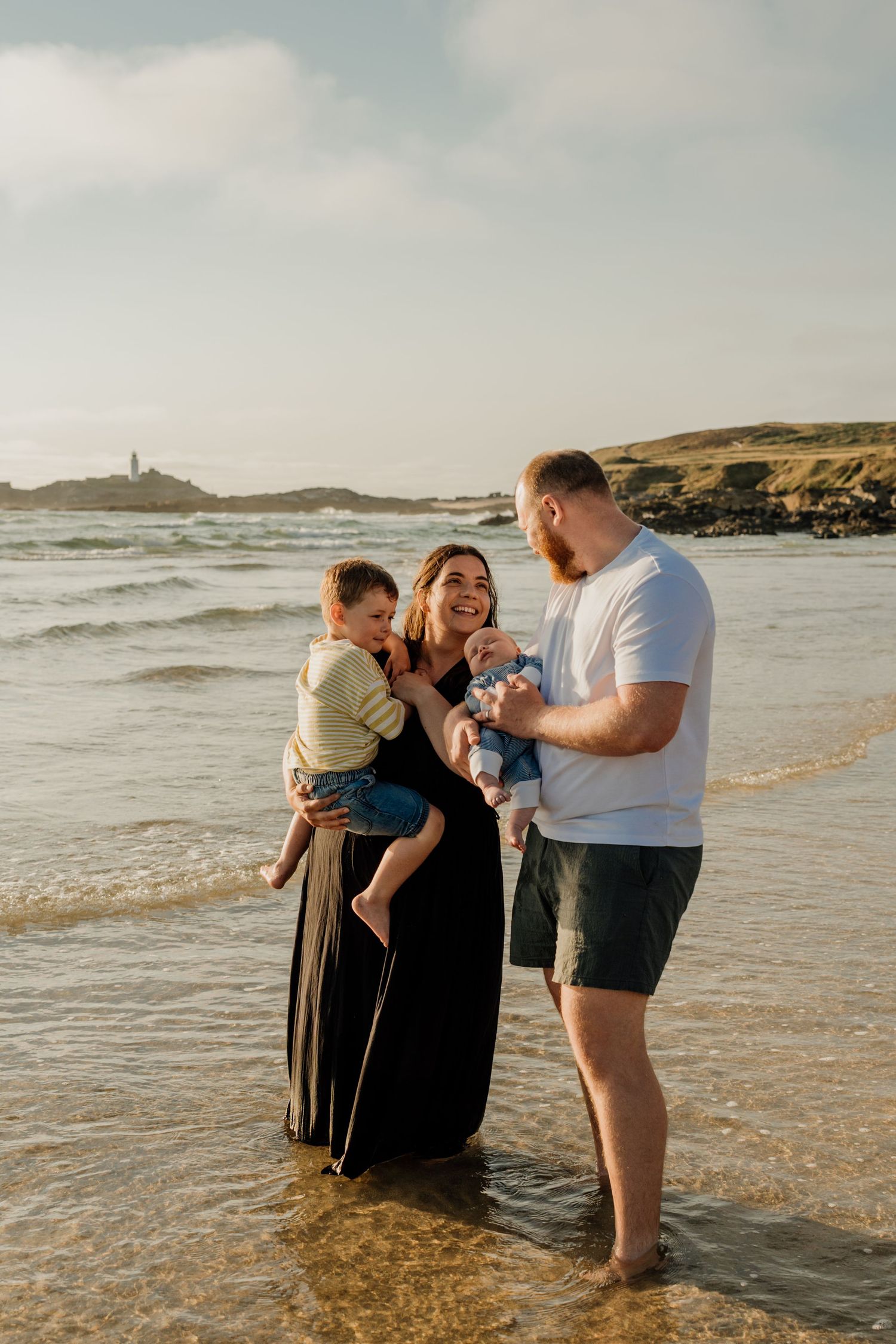 Family of four enjoying a sunny beach day, parents holding two young children near ocean waves with coastal hills in background.