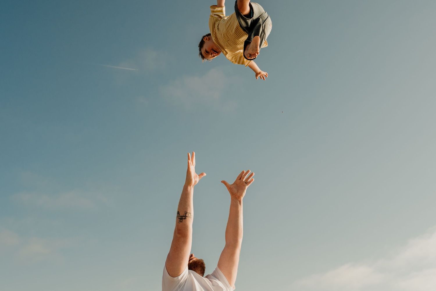 Adult tossing a child high into the blue sky outdoors, arms raised to catch the laughing child mid-air.