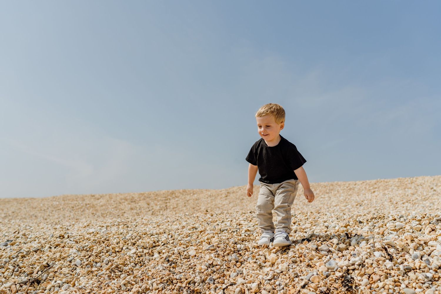 Child exploring and playing on a sunny pebble beach with blue skies overhead.
