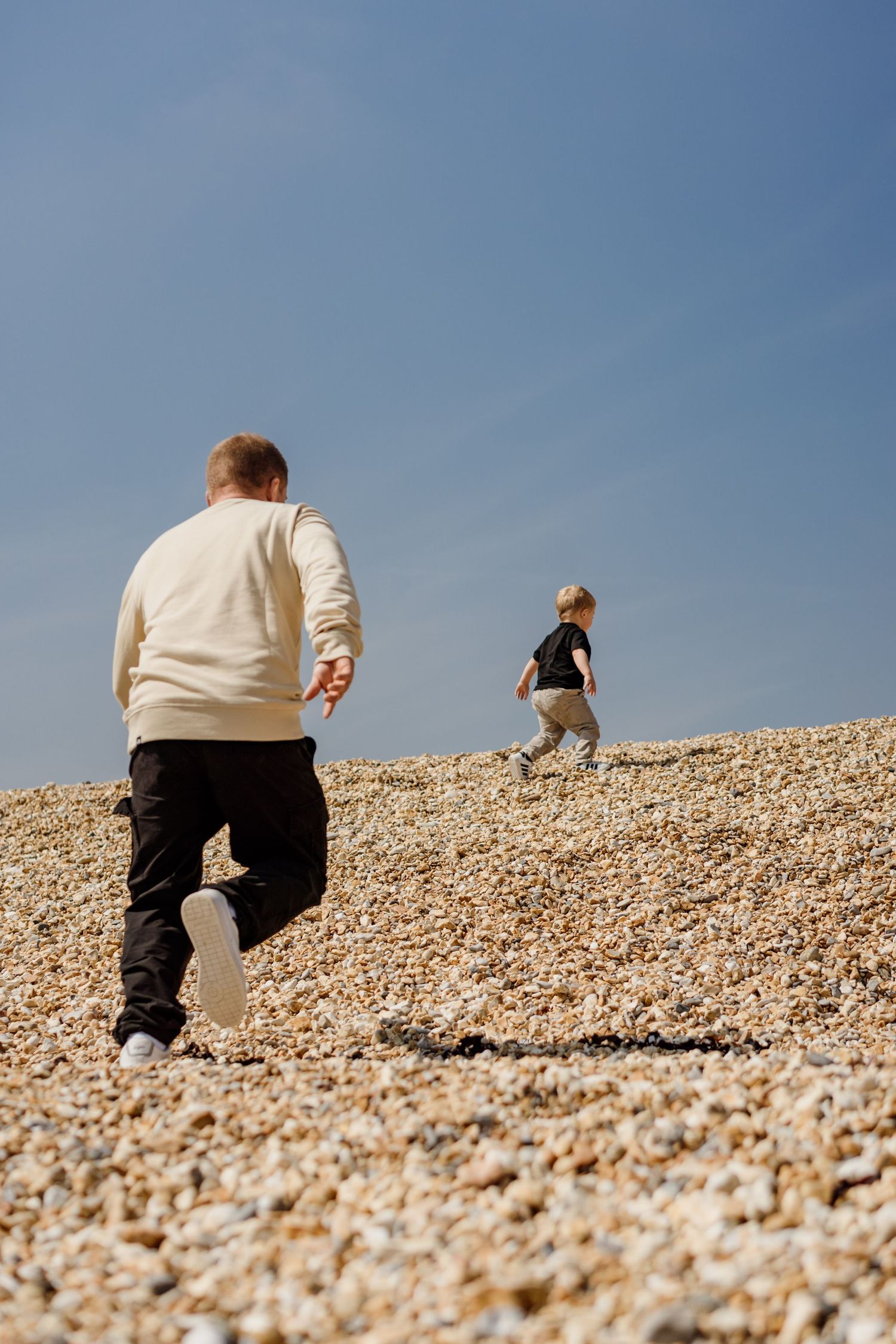 People climbing up a steep pebble beach slope against bright blue sky.