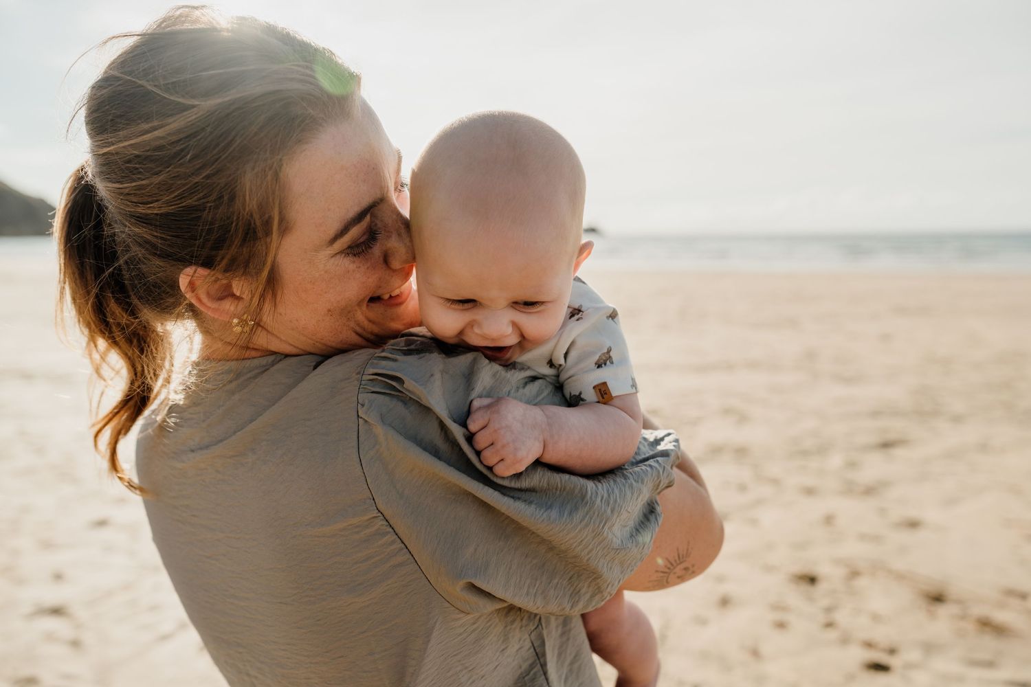 A young mother lovingly holds her smiling baby on a sunny beach, sharing a tender moment together.