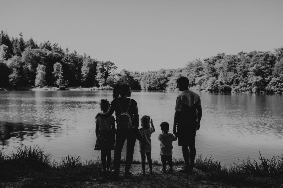 Silhouetted group gathering by a serene lake surrounded by trees in black and white.