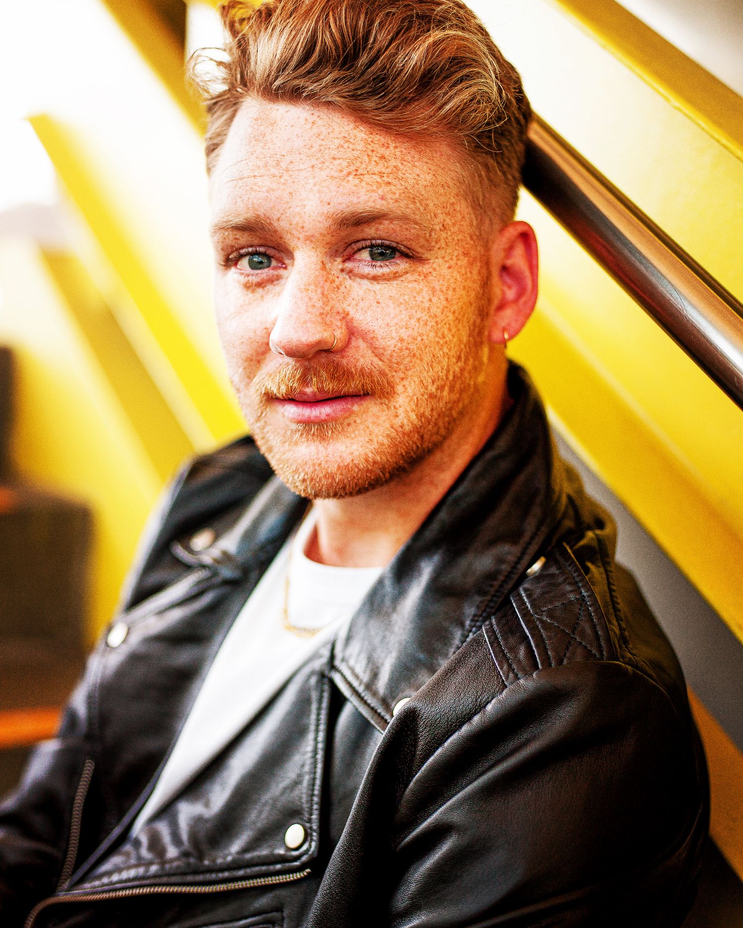 Portrait of a person with red hair and freckles wearing a black leather jacket against a yellow background.