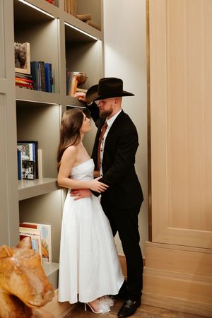 A romantic couple embraces in a library setting with built-in bookshelves, the woman wearing white and man in black.