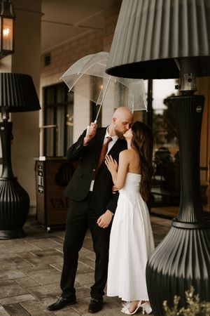 A couple shares an intimate moment under a clear umbrella on a covered walkway with classic columns.