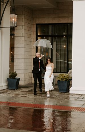 Wedding couple walking with clear umbrellas along a covered exterior building walkway in the rain.