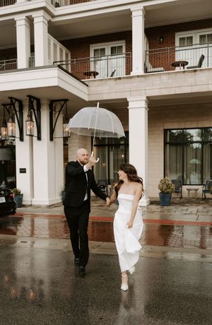 Wedding couple playfully walking in rain under clear umbrellas by columned building entrance.