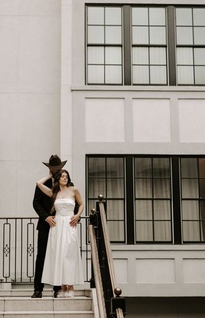 A couple shares a romantic moment on a white staircase with large windows in the background.