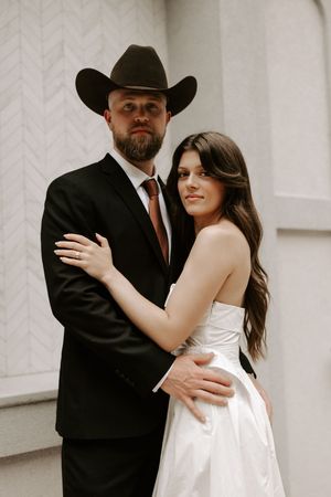 A couple in wedding attire share an intimate moment against a white wall, with the groom wearing a black cowboy hat.