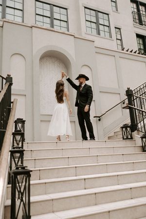 Bride in white dress and groom in black suit pose on architectural steps.