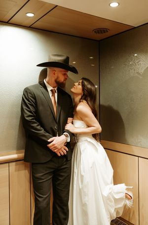A couple shares an intimate moment in a hallway, with the person wearing a cowboy hat and suit.