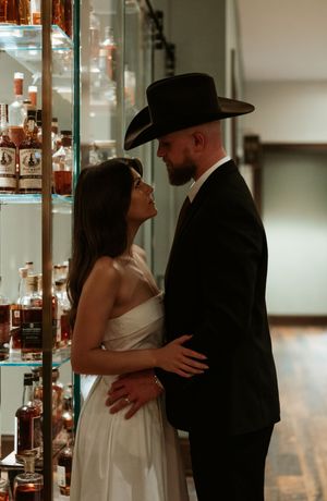 A romantic couple embraces near a display case, with the groom wearing a black cowboy hat and suit.