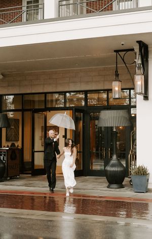 A couple shares a playful moment under clear umbrellas in front of a modern building entrance.