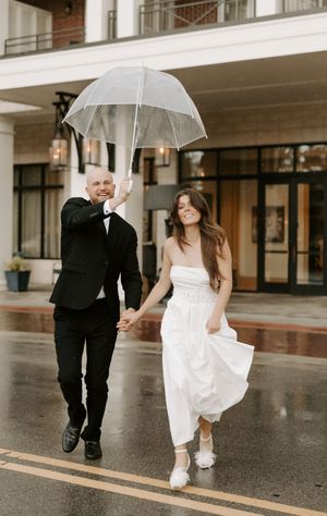 Two people dance joyfully in formal attire while holding clear umbrellas on wet pavement.