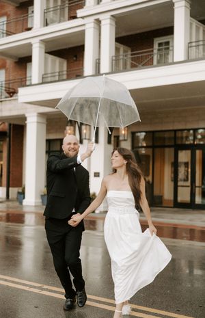 A couple in wedding attire dances with umbrellas on a rainy day outside a modern building.