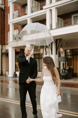 Two people share intimate moments while holding clear umbrellas outside a columned building.