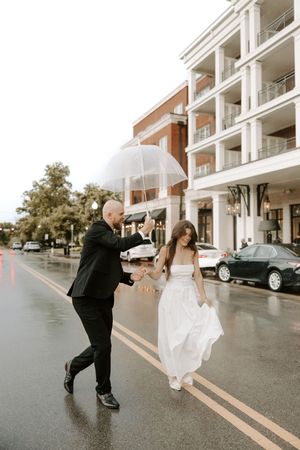 Wedding couple dances joyfully on a wet street in front of a modern apartment building.