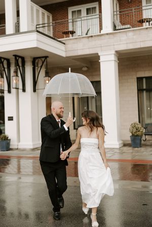 Newlyweds dance and twirl together on wet pavement outside a brick building.