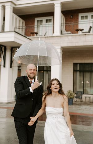 Wedding couple laughs and dances with a clear umbrella on a rainy afternoon.