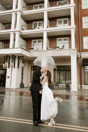 Wedding couple dances playfully in front of a modern apartment complex on a rainy day.