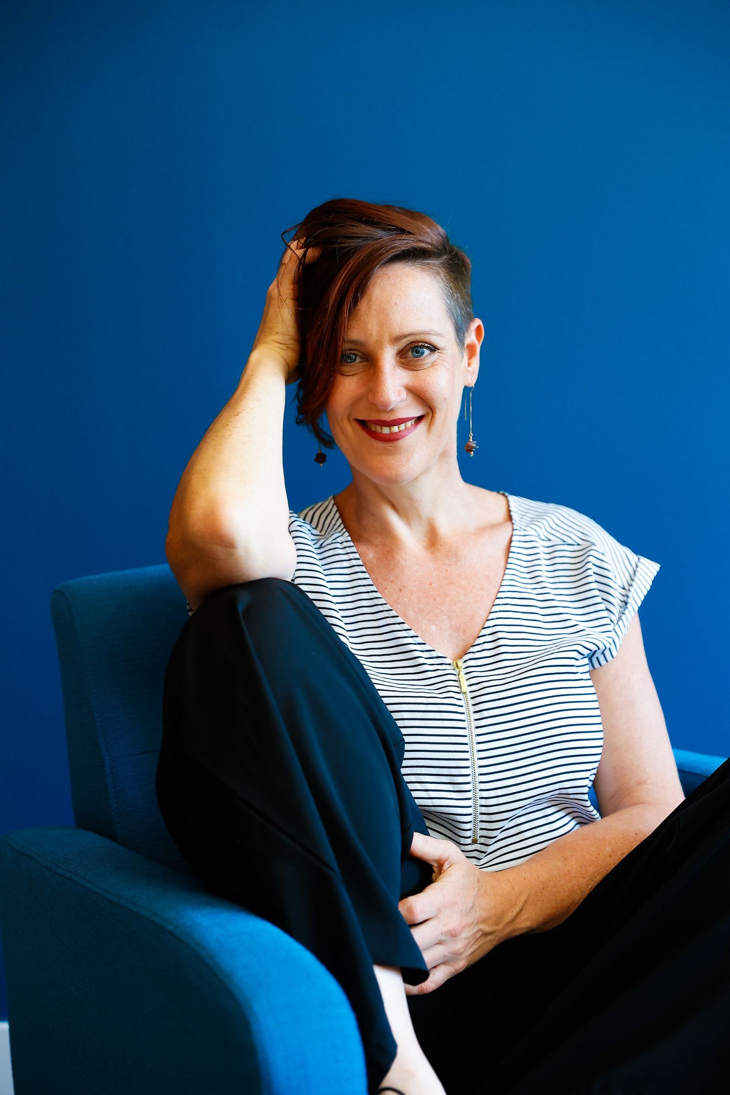 Someone in a striped top sits casually on a blue chair against a vibrant blue wall, smiling warmly.