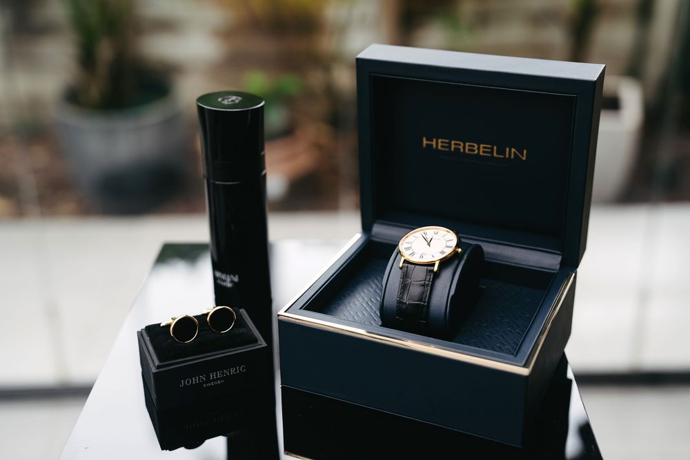 Luxury watch displayed in dark blue Hebrelin presentation box next to black cylindrical container on white surface.