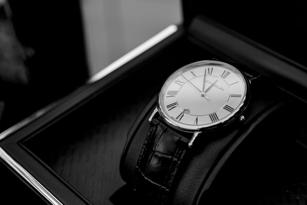 Elegant luxury watch with white Roman numeral face and black leather strap displayed in an open presentation box.