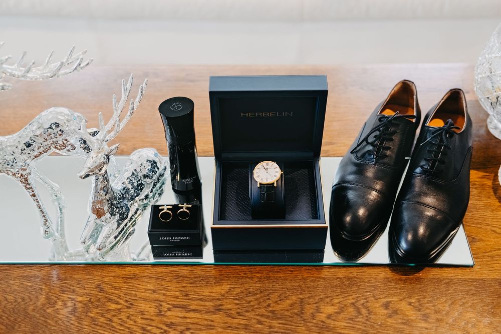 Black leather dress shoes, cologne bottle, and ring box arranged on wooden surface for wedding day preparation details.