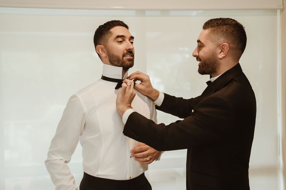 Man in a black suit jacket helps adjust a bowtie on someone wearing a white dress shirt against a light background.