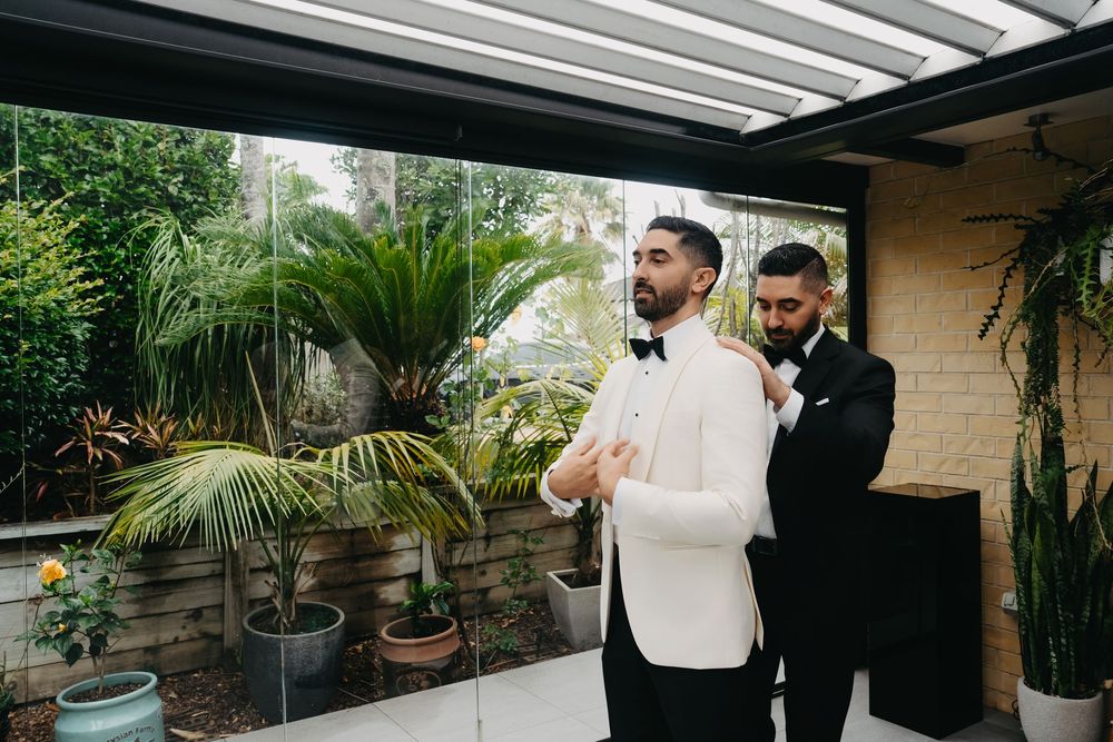 Two people in formal white and black tuxedos stand together on a patio with tropical plants in the background.