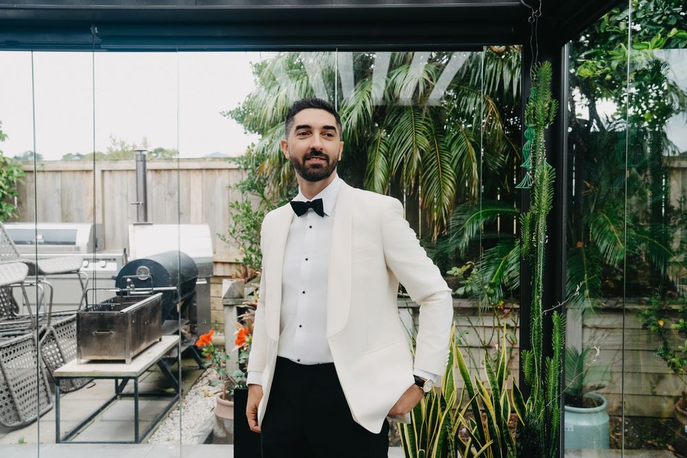 A person in formalwear poses in an outdoor garden setting with tropical plants and greenery.