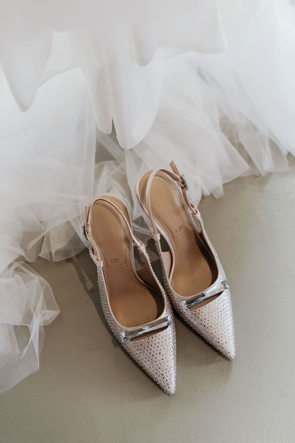 Elegant white pointed-toe slingback bridal shoes with rhinestone details against delicate tulle fabric background.