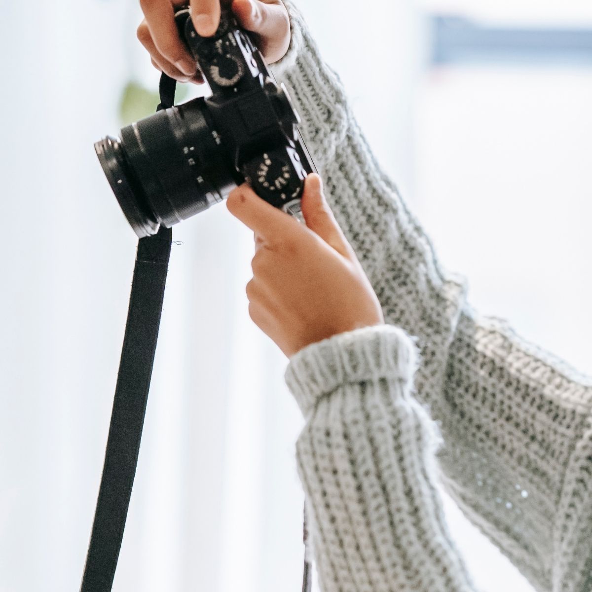 Hands wearing chunky gray knit sweater holding a black camera lens against a bright white background.