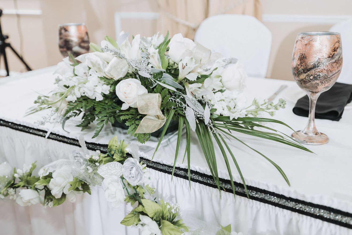Elegant white floral centerpiece with greenery arranged on a table next to a decorated wine glass at a wedding reception.