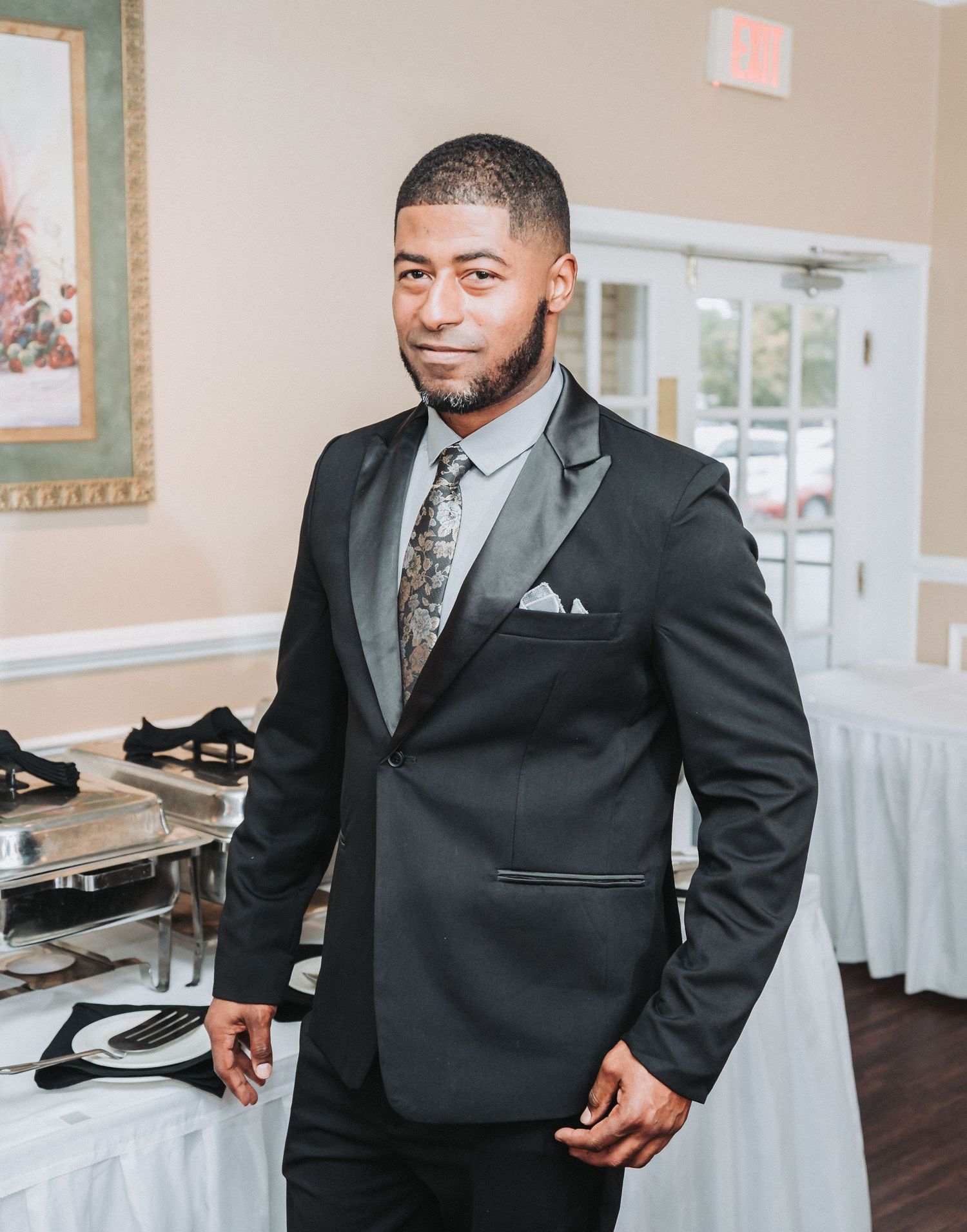 A well-dressed person in a dark suit and patterned tie stands in a formal event venue setting.