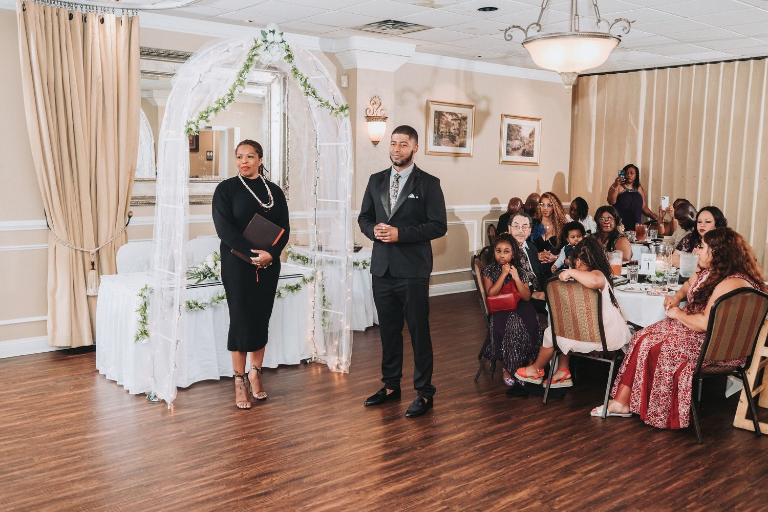 Indoor wedding ceremony setup with white floral arch and guests seated in elegant reception room.