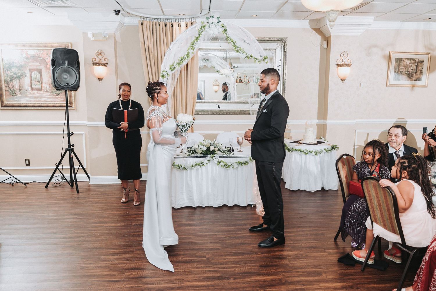 Wedding ceremony in a decorated banquet hall with white floral arch and guests watching the proceedings.