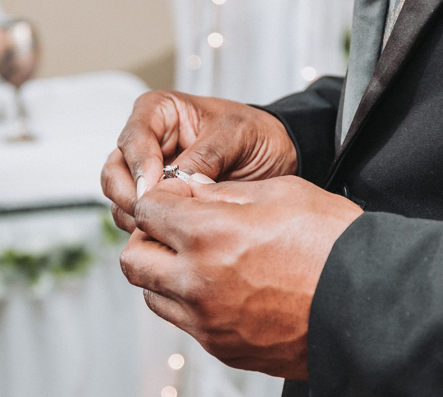 Close-up view of hands in a formal suit adjusting wedding ring during a ceremony with soft bokeh lights in background.