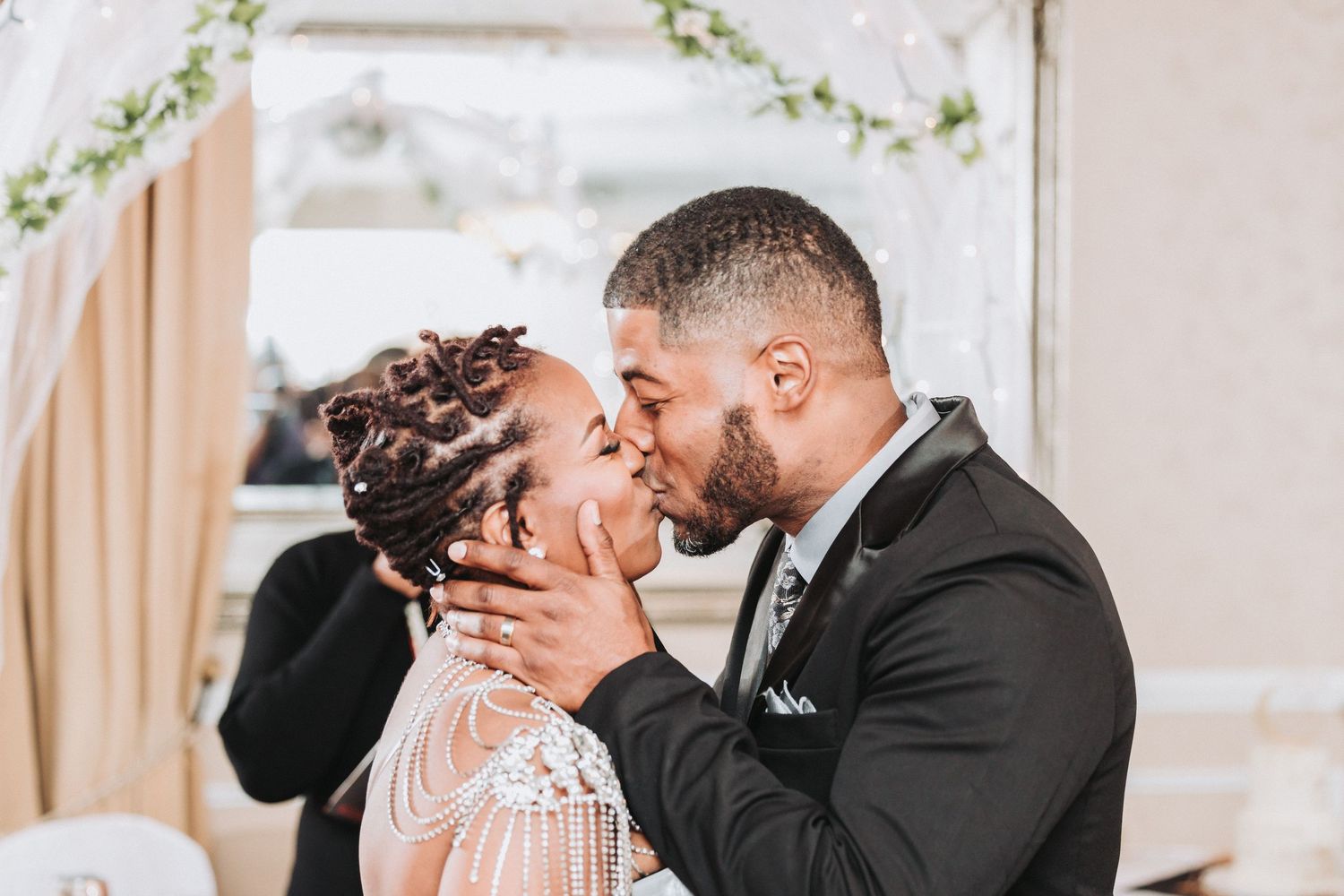 Two people share a romantic kiss during what appears to be a wedding ceremony with floral decorations.