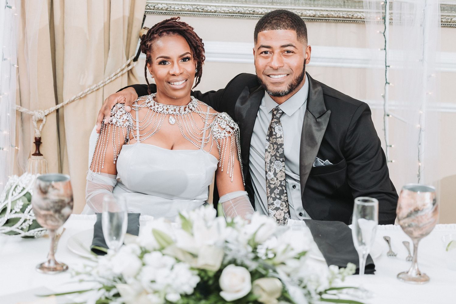 Newlywed couple smiling together at their wedding reception with white floral centerpiece on table.