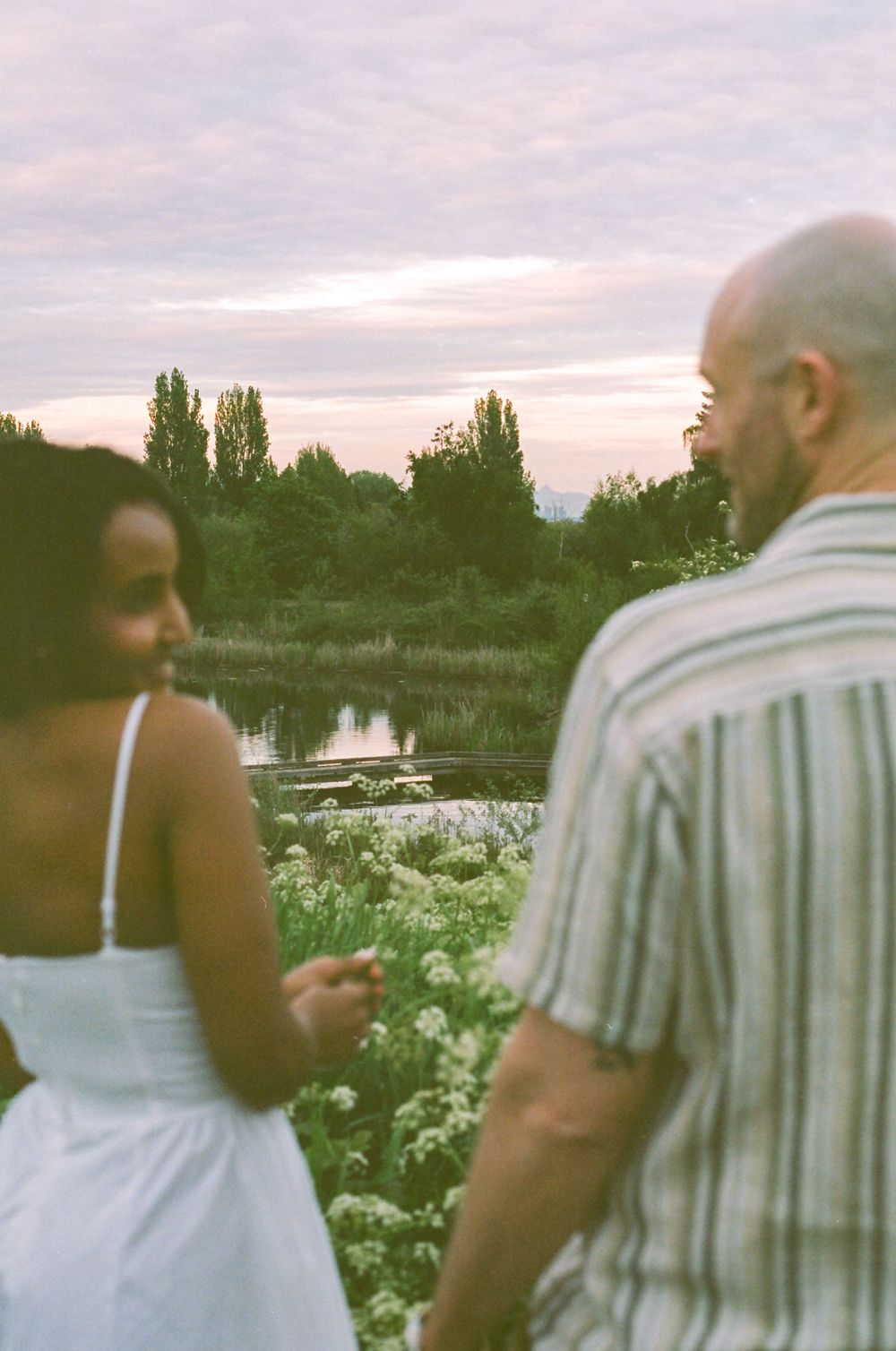 Couple standing together among white wildflowers at dusk near a lake.