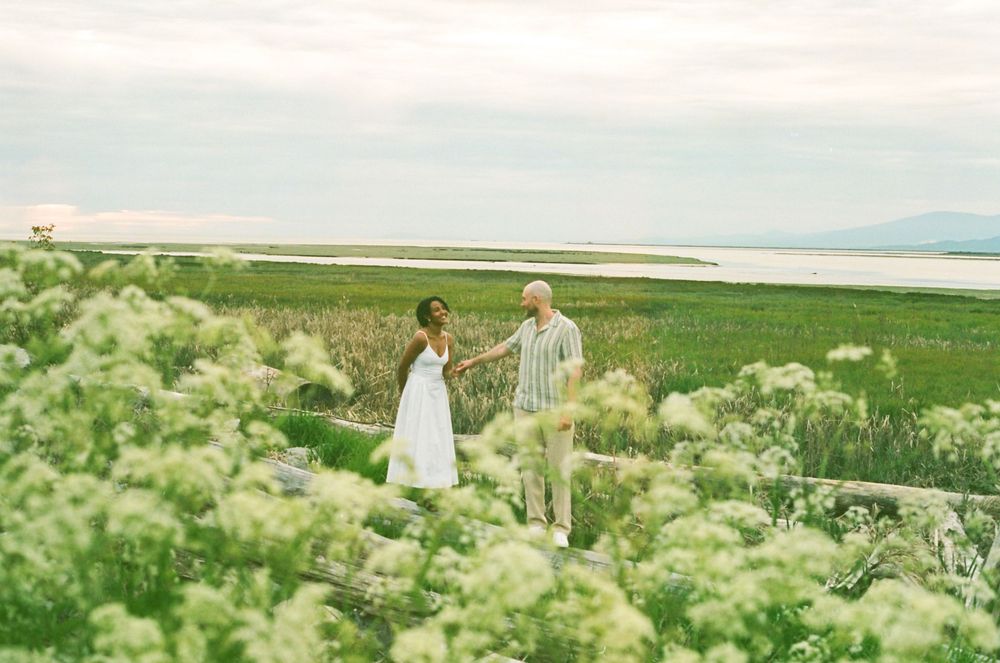 A romantic couple poses together in a scenic field filled with blooming white wildflowers on a sunny day.