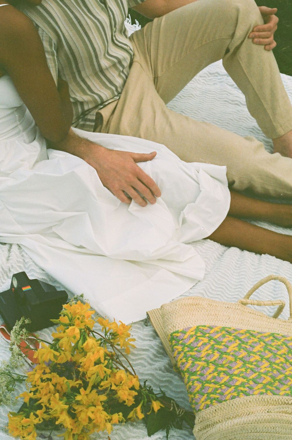 Close-up of white dress and striped shirt with yellow flowers and a woven bag on picnic blanket.