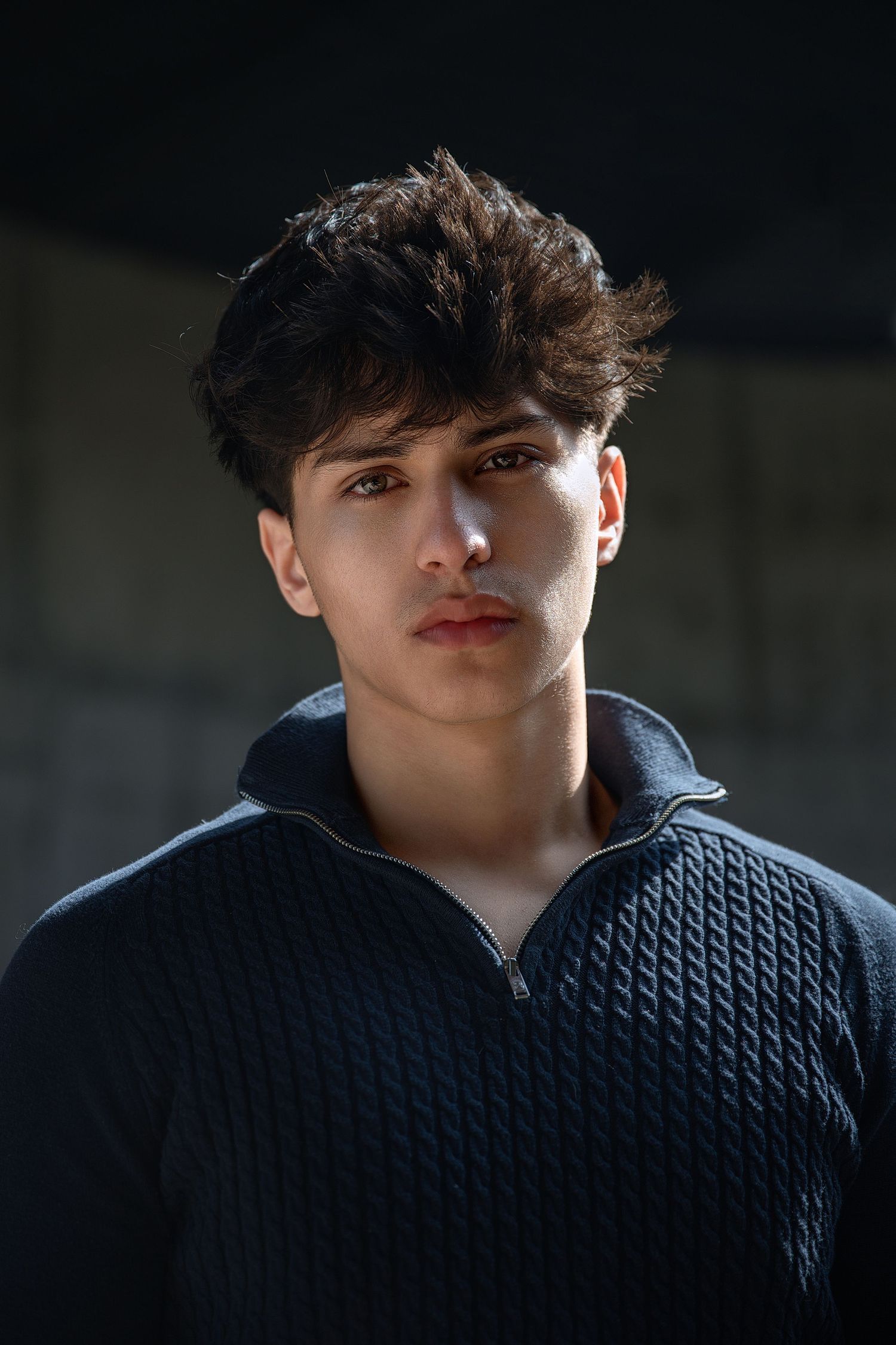 Portrait of a young person with dark styled hair wearing a black quilted zip-up sweater in dramatic lighting.